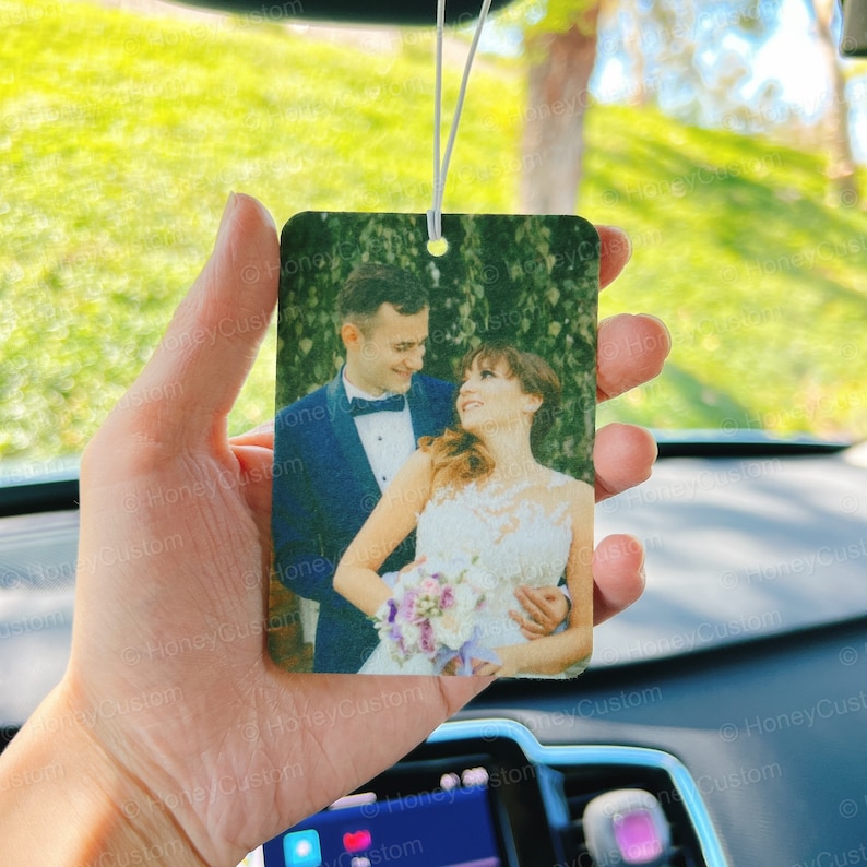May include: A white car air freshener with a photo of a couple in wedding attire. The couple is smiling and holding a bouquet of flowers. The air freshener is hanging from the rearview mirror.