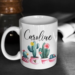 May include: White ceramic mug with a black cursive name "Caroline" and a design of potted cacti with pink and yellow flowers.