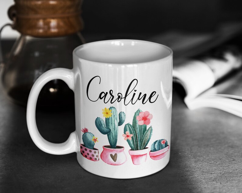 May include: White ceramic mug with a black cursive name "Caroline" and a design of potted cacti with pink and yellow flowers.