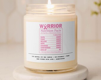 May include: A white soy wax candle with a pink ribbon and the text "WARRIOR Nutrition Facts" on the label. The label lists "Faith, Fight, Believe, Hope, Courage, Confidence, Give Up" with a percentage value of 1000% for each except "Give Up" which is 0%. The candle is on a white marble coaster.