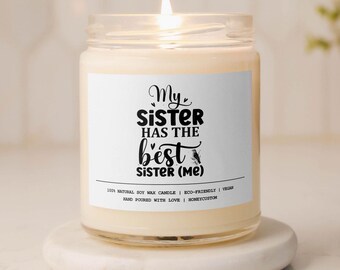 May include: A white soy wax candle in a clear glass jar with a black and white label that reads "My Sister Has The Best Sister (Me)". The candle is lit and the label also includes the text "100% Natural Soy Wax Candle | Eco-Friendly | Vegan | Hand Poured With Love | Honeycustom".