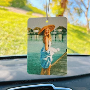 May include: A woman wearing a white dress and a straw hat stands on a wooden dock over the water. The water is blue and there are buildings in the background.