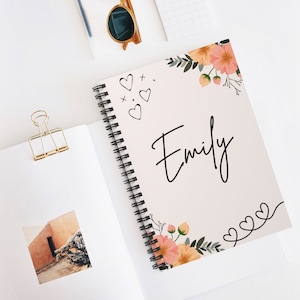 May include: A spiral-bound notebook with the name "Emily" in black script. The cover is light pink with floral and heart designs. Sunglasses and a gold paper clip are also visible.
