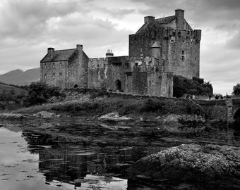 May include: Black and white photograph of a medieval castle on a small island, with a bridge leading to the entrance. The castle is reflected in the water, under a cloudy sky. The image is a landscape shot.