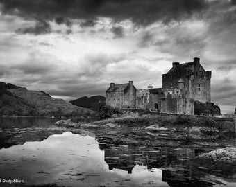 May include: Black and white photograph of Eilean Donan Castle in Scotland. The medieval castle sits on an island at the point where three sea lochs meet. The castle's reflection is visible in the water. The sky is overcast.