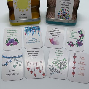 May include: A set of 10 affirmation cards with colorful illustrations and positive messages. The cards are in a small tin box with a yellow lid and a blue lid.