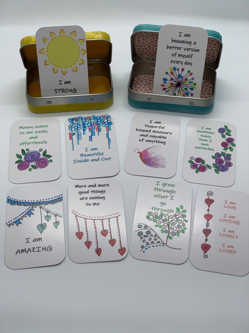 May include: A set of 10 affirmation cards with colorful illustrations and positive messages. The cards are in a small tin box with a yellow lid and a blue lid.