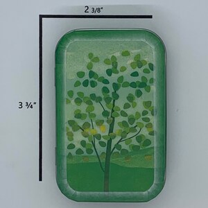 May include: A green metal tin with a rounded rectangular shape. The tin features a design of a green tree with yellow leaves against a light blue sky. The tin measures 2 3/8 inches by 3 3/4 inches.