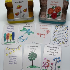 May include: A set of affirmation cards in a small tin box. The cards feature positive messages such as "I am abundant", "I am grateful", "I am free of limiting beliefs", and "Everything is always working out for me".