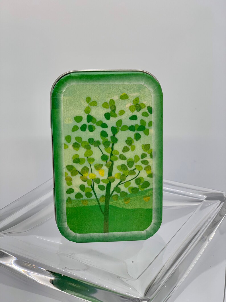 May include: A small, rectangular tin with a green and white design featuring a stylized tree with green leaves and a brown trunk. The tin has a silver rim.