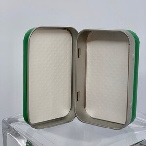 May include: A green and silver metal box with a white foam interior. The box is open and the foam is visible.