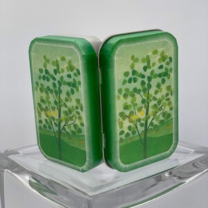 May include: A small, green, hinged tin box with a white interior. The box is decorated with a stylized image of a tree with green leaves and yellow flowers. The box is closed and standing upright on a clear glass surface.