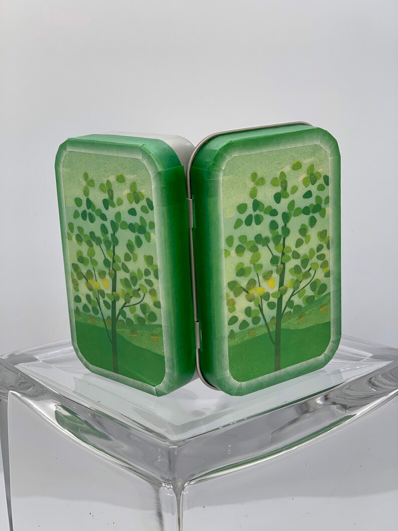 May include: A small, green, hinged tin box with a white interior. The box is decorated with a stylized image of a tree with green leaves and yellow flowers. The box is closed and standing upright on a clear glass surface.