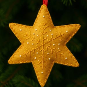 May include: A mustard yellow, six-pointed star ornament with a red ribbon for hanging. The star is embroidered with a snowflake design and dotted with small white beads. The ornament is hanging in front of a green, blurred background.
