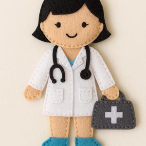 Op de afbeelding: Vilten dokterfiguur met zwart haar, een witte jas en een stethoscoop. De figuur houdt een grijze medische tas met een wit kruis vast. De dokter draagt blauwe schoenen.