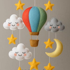 May include: A felt baby mobile featuring a hot air balloon in red, blue, and green, with smiling white clouds, yellow stars, and a yellow crescent moon. The text "Instant Download" and "PDF tutorial + Patterns" are visible.