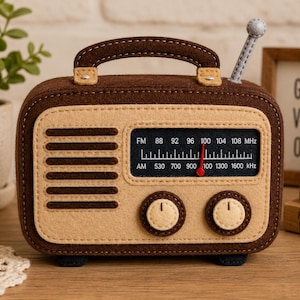 May include: A brown and beige felt radio with a top handle and a grey antenna. The front features a speaker grille, two circular knobs, and a black display panel showing FM and AM frequencies with a red needle pointing to 100 MHz.