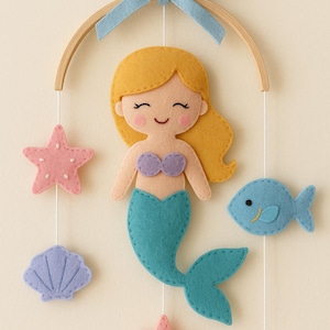 May include: A felt mermaid mobile with a light blue bow, wooden hoop, and hanging felt sea creatures. The mermaid has yellow hair, a purple top, and a teal tail. Includes a pink starfish, a blue fish, and a purple seashell.