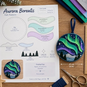 May include: A printed Aurora Borealis felt pattern sheet with cutting and sewing guides, alongside a finished circular felt ornament featuring green and purple waves, mountains, and trees. A wooden ruler shows a 10 cm or 4 inch diameter.