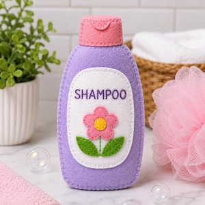 Puede incluir: Una botella de champú de fieltro hecha a mano en color morado claro con una tapa rosa. La parte frontal presenta una etiqueta ovalada blanca con la palabra SHAMPOO bordada en morado, acompañada de una flor rosa con centro amarillo y dos hojas verdes.