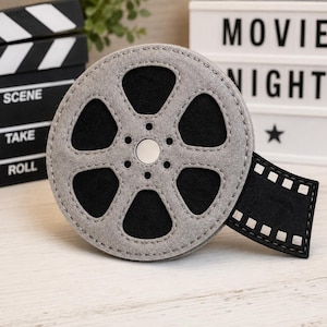Può includere: Un ornamento circolare in feltro grigio a forma di bobina cinematografica con ritagli neri e un bottone argentato centrale. Una corta striscia di pellicola in feltro nero con perforazioni quadrate bianche sporge dal lato destro.