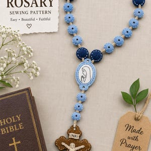 May include: A handmade felt rosary featuring light blue and dark blue circular beads, a central oval pendant with a white illustration of a person in prayer, and a brown cross with a white figure of a person and the text INRI at the top.