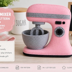Puede incluir: Una batidora de pie de fieltro hecha a mano en rosa claro y gris, con un bol mezclador gris extraíble y un accesorio de varillas. La batidora tiene un dial de control circular, una pequeña pantalla rectangular negra y costuras visibles.