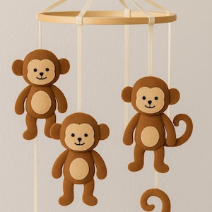 Può includere: Mobile per bambini con tre figure di scimmia in feltro marrone, ciascuna con pancia beige e viso sorridente. Le scimmie sono sospese a un anello di legno con nastri color crema. Ideale per una cameretta.