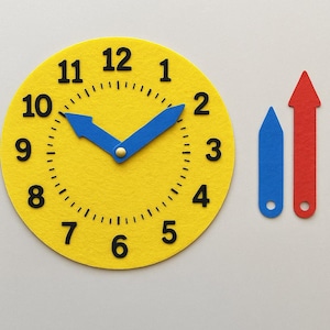 Puede incluir: Un reloj de fieltro amarillo con números negros y manecillas azules que señalan las 10:10. Dos formas de fieltro adicionales: una flecha azul y una flecha roja, están junto al reloj. El reloj es una herramienta de aprendizaje.