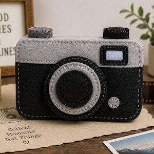 May include: A handcrafted felt camera toy featuring a black body and a light grey top section. The design includes a circular black lens with grey trim, a small rectangular light blue viewfinder, and two grey and black circular buttons on top.