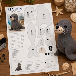 May include: A sea lion felt pattern document featuring numbered cut-out shapes, assembly instructions, and a finished dark gray felt sea lion plush toy with a beige muzzle, black nose, and black eyes, displayed alongside sewing supplies.