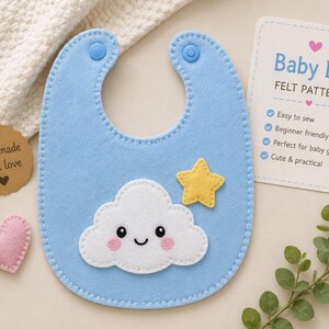 Puede incluir: Un babero de fieltro azul claro para bebé con una aplicación de nube blanca sonriente con mejillas rosadas y una estrella amarilla. El babero tiene dos broches de presión de plástico azul. Una tarjeta Baby Bib FELT PATTERN y una etiqueta handmade with love.