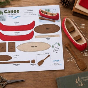 May include: A felt canoe sewing pattern sheet with printed instructions, diagrams, and labels. Beside it sits a finished red and beige felt canoe with a brown paddle, measuring 6 inches long, 1.6 inches wide, and 1.6 inches high, with a 4.7 inch paddle.