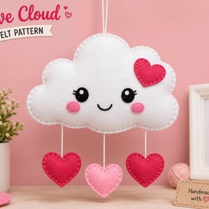 May include: A white felt cloud ornament with a smiling face, pink cheeks, and a dark pink heart on its top right. Three dangling hearts in shades of pink hang from the cloud by white strings. The text Love Cloud FELT PATTERN is visible.