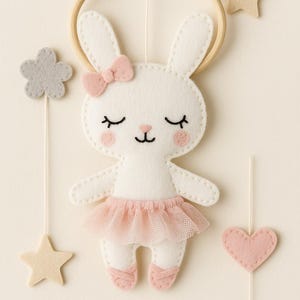 Könnte beinhalten: Ein weißes Filz-Hase-Mobile mit rosa Schleife, Tutu und Ballettschuhen. Das Mobile enthält einen Holzreifen, Stern- und Blumenakzente und ein Herz. Der Hase hat geschlossene Augen und ist an einer Schnur aufgehängt.
