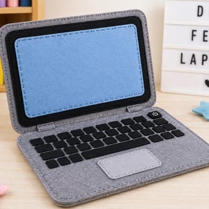 Op de afbeelding: Een laptop-speeltje van grijze vilt met een lichtblauw scherm, een zwart toetsenbord met individuele zwarte toetsen, een aan-uitknop icoon en een lichtgrijs trackpad. De laptop is afgewerkt met zichtbare stiksels langs de randen van het vilt.