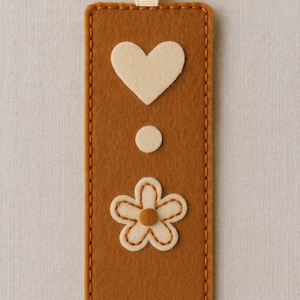 May include: A brown felt bookmark with a cream-colored heart, dot, and flower design. The bookmark has a cream-colored ribbon and is stitched with orange thread. A decorative item for books.