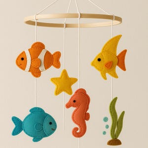 May include: A handmade baby mobile featuring felt sea creatures. The mobile includes an orange clownfish, yellow fish, blue fish, orange seahorse, yellow starfish, and green seaweed, all suspended from a wooden hoop.