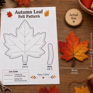 Puede incluir: Una hoja de papel blanca titulada Autumn Leaf Felt Pattern con dibujos de líneas negras de una hoja y un tallo, una guía de líneas y el texto Happy Crafting. Al lado hay una hoja de fieltro terminada de 11 cm por 10 cm en tonos rojos.