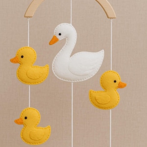 May include: A baby mobile featuring a white swan and three yellow ducklings suspended from a wooden arch. The swan and ducks are made of felt and have orange beaks and black eyes. The mobile is designed for a nursery.