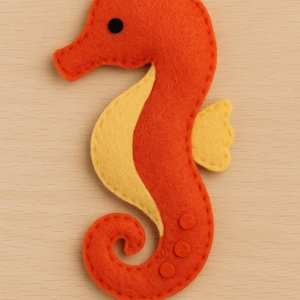 May include: A handmade felt seahorse in orange and yellow. The seahorse has a curved body, a small head, and a curled tail. The seahorse has a yellow belly and a small yellow fin. The seahorse has three small orange circles on its body and a black eye.