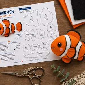 May include: A clownfish felt sewing pattern sheet with printed templates, assembly notes, and a materials list. The document features a finished orange and white clownfish plush, alongside a pair of metal scissors and craft supplies.