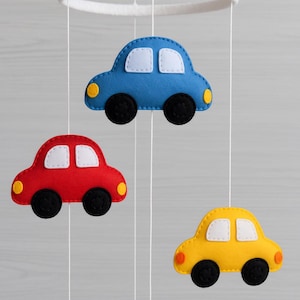 May include: A baby mobile with three felt cars in blue, red, and yellow. Each car has black wheels, white window details, and yellow or orange headlights. The cars are suspended from a white ring by thin white strings.