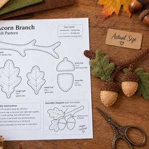 May include: A white paper pattern for an acorn branch craft project with black text and diagrams, alongside a finished felt acorn branch, metal scissors, and a wooden ruler showing measurements in centimetres on a wooden surface.