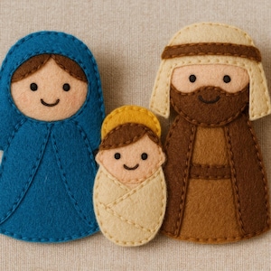 Pode incluir: Um presépio de feltro com três figuras: Maria com uma túnica azul, José com uma túnica castanha e o menino Jesus envolto numa manta creme. Cada figura tem um rosto sorridente e é costurada à mão.