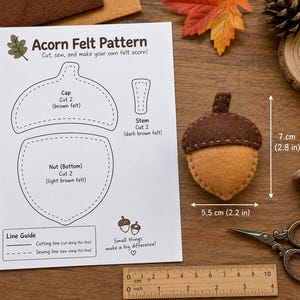 Può includere: Un foglio di carta bianco intitolato Acorn Felt Pattern con disegni a linee nere per un cappuccio, un gambo e una noce, accanto a una ghianda in feltro marrone e beige che misura 7 cm per 5,5 cm, con un righello in legno da 10 cm.