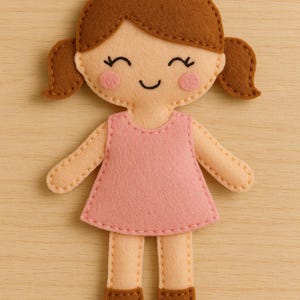 Könnte beinhalten: Eine handgefertigte Filzpuppe mit braunen Zöpfen, rosigen Wangen und einem lächelnden Gesicht. Die Puppe trägt ein rosa Kleid und hat beige Arme und Beine mit braunen Schuhen. Die Puppe ist mit einem Kontrastfaden zusammengenäht.