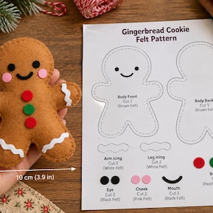 Pode incluir: Um boneco de gengibre de feltro castanho com 12 cm por 10 cm, com olhos pretos, boca preta, bochechas cor-de-rosa e três botões vermelhos e verdes. Ao lado, encontra-se uma folha impressa com o molde de feltro para bolachas de gengibre.