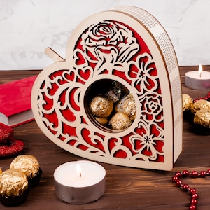May include: A wooden heart-shaped box with a red floral design and a cutout window revealing a collection of chocolate candies wrapped in gold foil. The box is sitting on a wooden surface with red beads and candles.