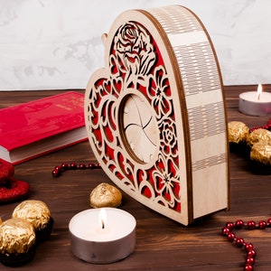 May include: A wooden heart-shaped box with a red floral design and a cutout in the center. The box is surrounded by red beads, gold-wrapped chocolates, and two white candles.